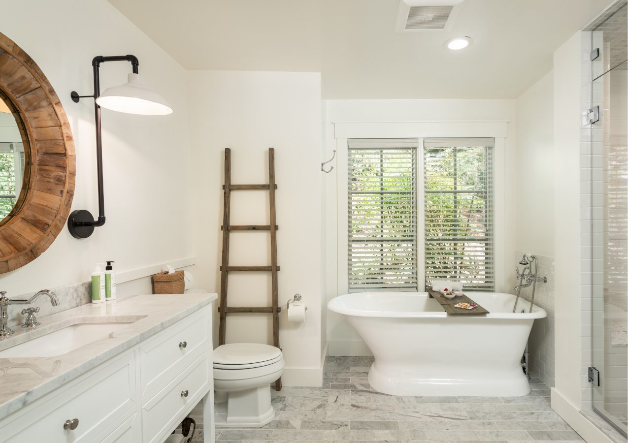 Interior of Cottage Room Bathroom: white walls, sink and counter to left with drawers and round mirror hung above, elegant stand-alone bathtub in back with vertical windows above, white toilet with ladder decoration on wall, glass door for shower on right.