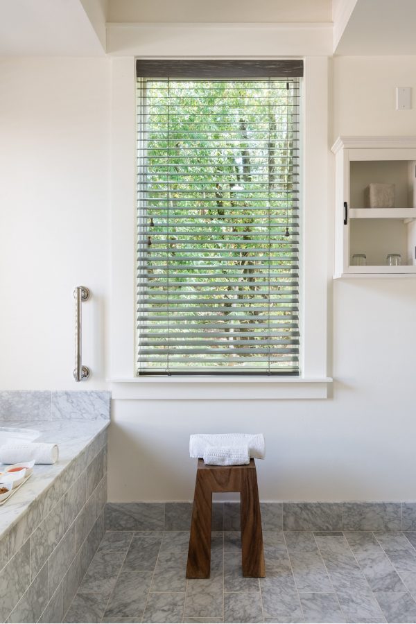 interior of large, elegant bathroom: gray tiled bathtub, matching tiled floors, large vertical glass window, elegant sink and mirror to the right. looking out to greenery,