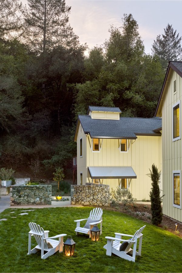 birdseye view of the Farmhouse back patio near the barn rooms at dusk. Set of three patio chairs in a circle with laterns, and backlit yellow barnhouse buildings