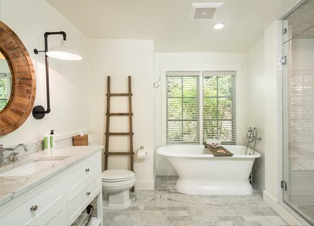 Interior of Cottage Room Bathroom: white walls, sink and counter to left with drawers and round mirror hung above, elegant stand-alone bathtub in back with vertical windows above, white toilet with ladder decoration on wall, glass door for shower on right.