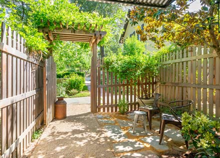 Exterior view of Cottage Room patio: tall wood picket fence with path out of a gate and lush greenery in the distance.