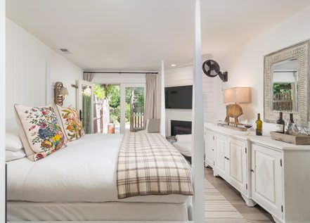 Interior of Cottage Room: side view of large bed with white bed posts and white bedding with plaid folded blanket at foot of bed. Counter-height dresser on other side of room with lamp and mirror hung above. Glass doors in back leading to patio with lush greenery.