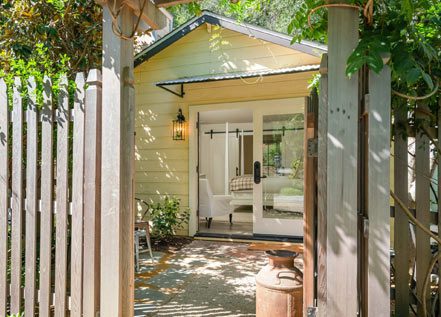 Exterior view of Cottage Room: looking through gate of wood picket fence, yellow exterior of building, beautiful glass double doors. Bed, wood floor and white chair are visible through glass doors.