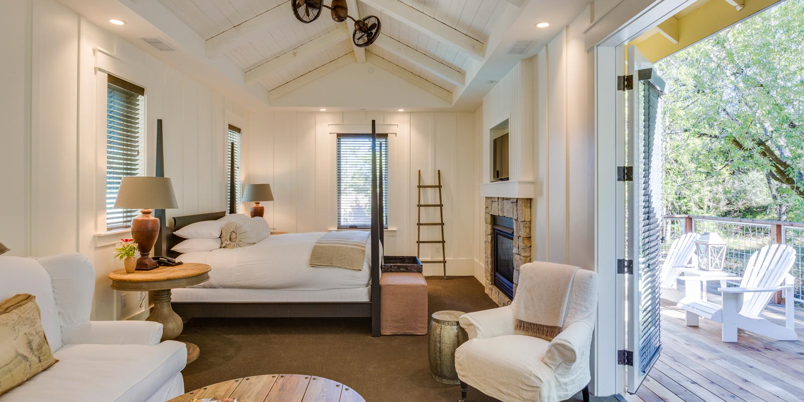 interior of Barn Junior Suite with ceiling fan, large bed with white bedding, white chair and love seat. porch door is open to a wood deck with white patio chairs