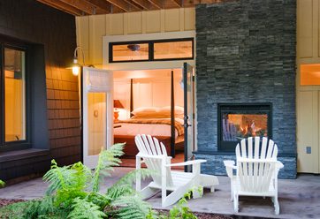 porch view of Barn Junior Suite featuring indoor, outdoor fireplace, white patio wood chairs, and a view inside room with large bed with white bedding