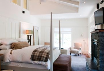 Interior view of Barn Junior Suite. White walls, stone fireplace with mounted TV above, large bed with white bedding with gray bedposts, high ceilings with ceiling fan.
