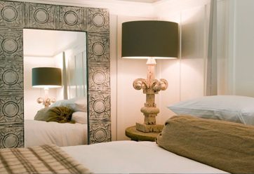 Interior close-up of Barn Junior Suite. Large bed with white bedding, tall, ornate lamb on night stand, large wall mirror with decorated frame.