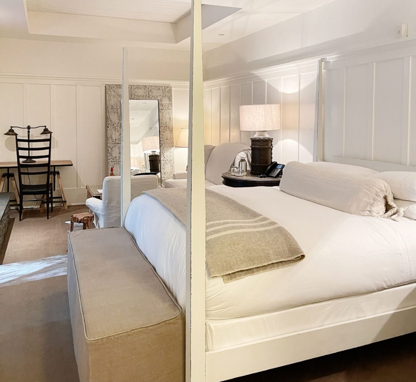 Interior of room with large feather bed with white bedding and white wood bed posts. Cushioned chairs and large lamp on night stand in background.