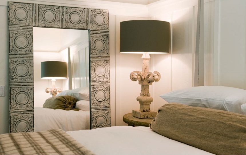 Interior close-up of Barn Junior Suite. Large bed with white bedding, tall, ornate lamb on night stand, large wall mirror with decorated frame.