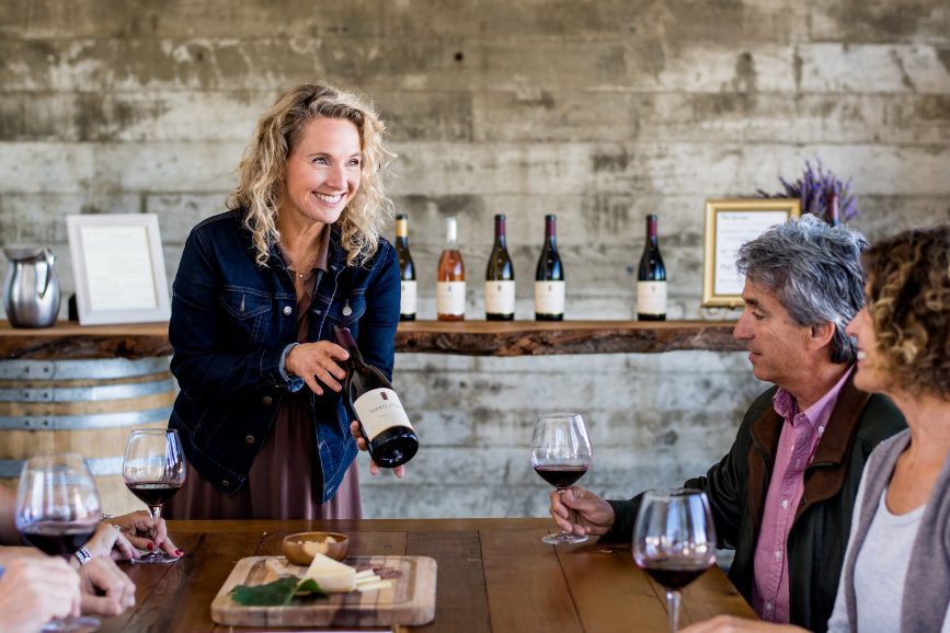 Farmhouse guests enjoying a tasting led by a sommelier in a private tasting room. Sommelier holding out bottle of wine to guests