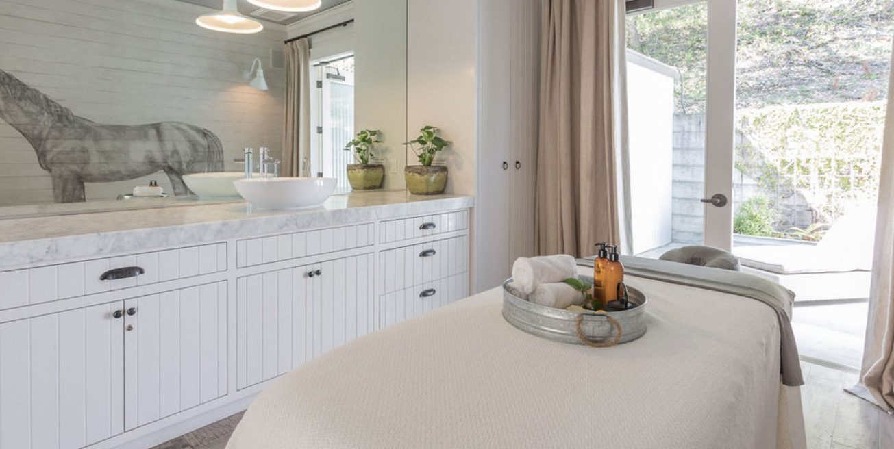 Spa room with massage table with tray of massage products, white wood cabinetry with sink and full wall mirror, glass door with a view of a patio.