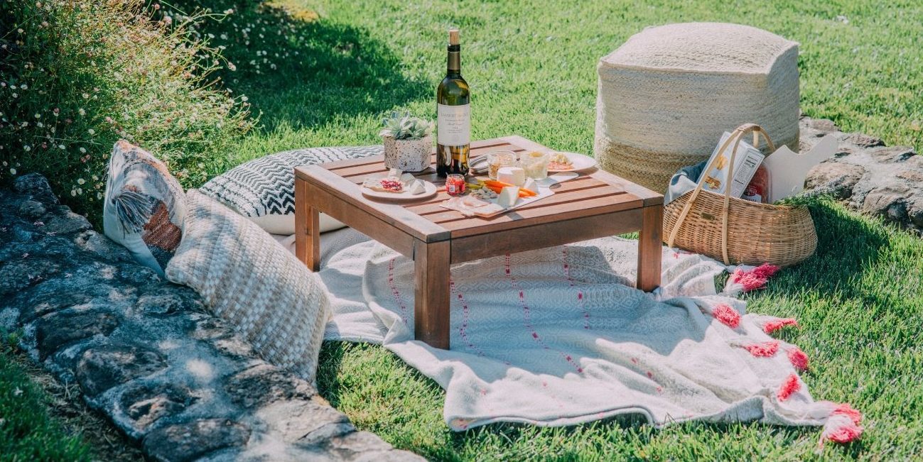 outdoor picnic: blankets and pillows on grass, picnic basket, and table with a bottle of wine and snacks