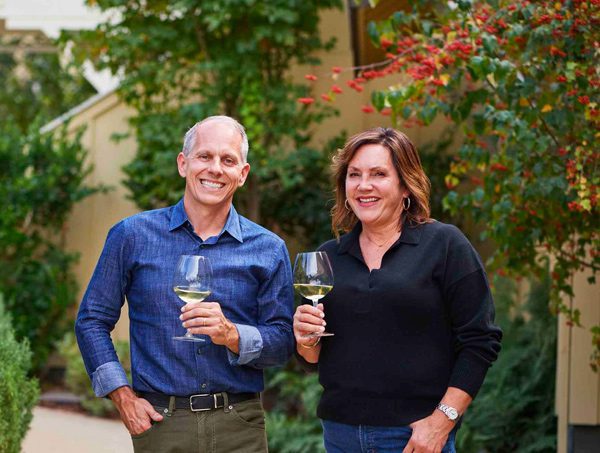 Photo of Joe and Catherine Bartolomei each holding a glass of wine smiling on the property of Farmhouse Inn