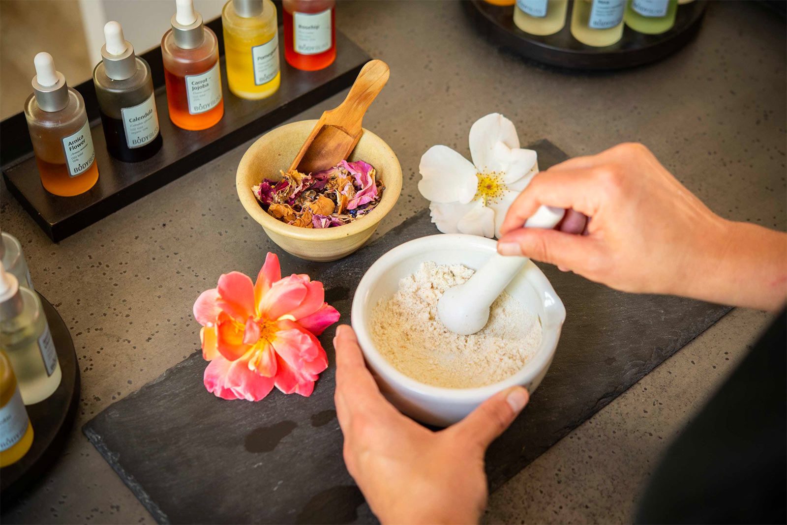 closeup of hands mixing a fine powder in a mortar and pestle. Two flowers and some bottles are in view.