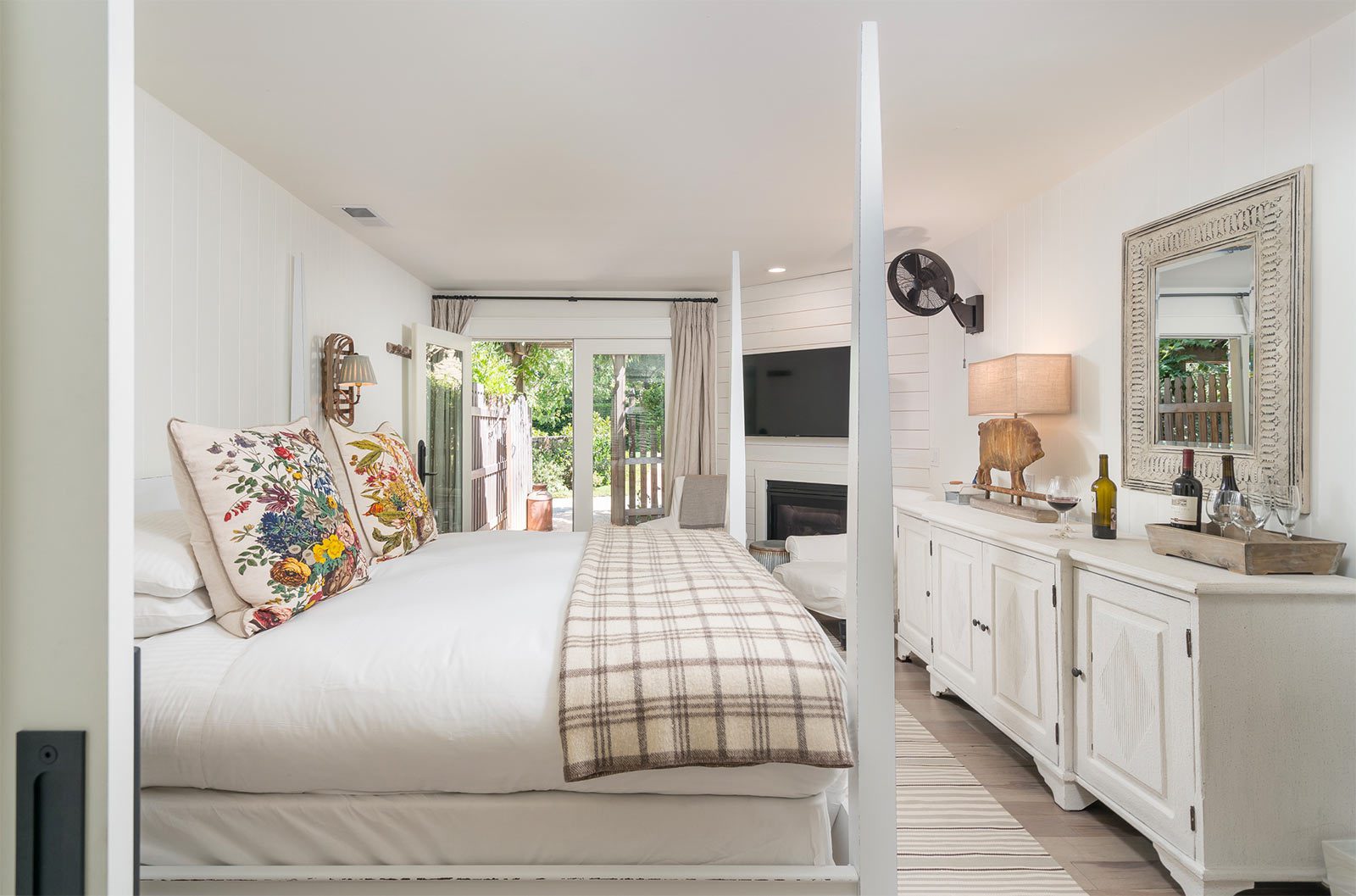 Interior of Cottage Room: side view of large bed with white bed posts and white bedding with plaid folded blanket at foot of bed. Counter-height dresser on other side of room with lamp and mirror hung above. Glass doors in back leading to patio with lush greenery.