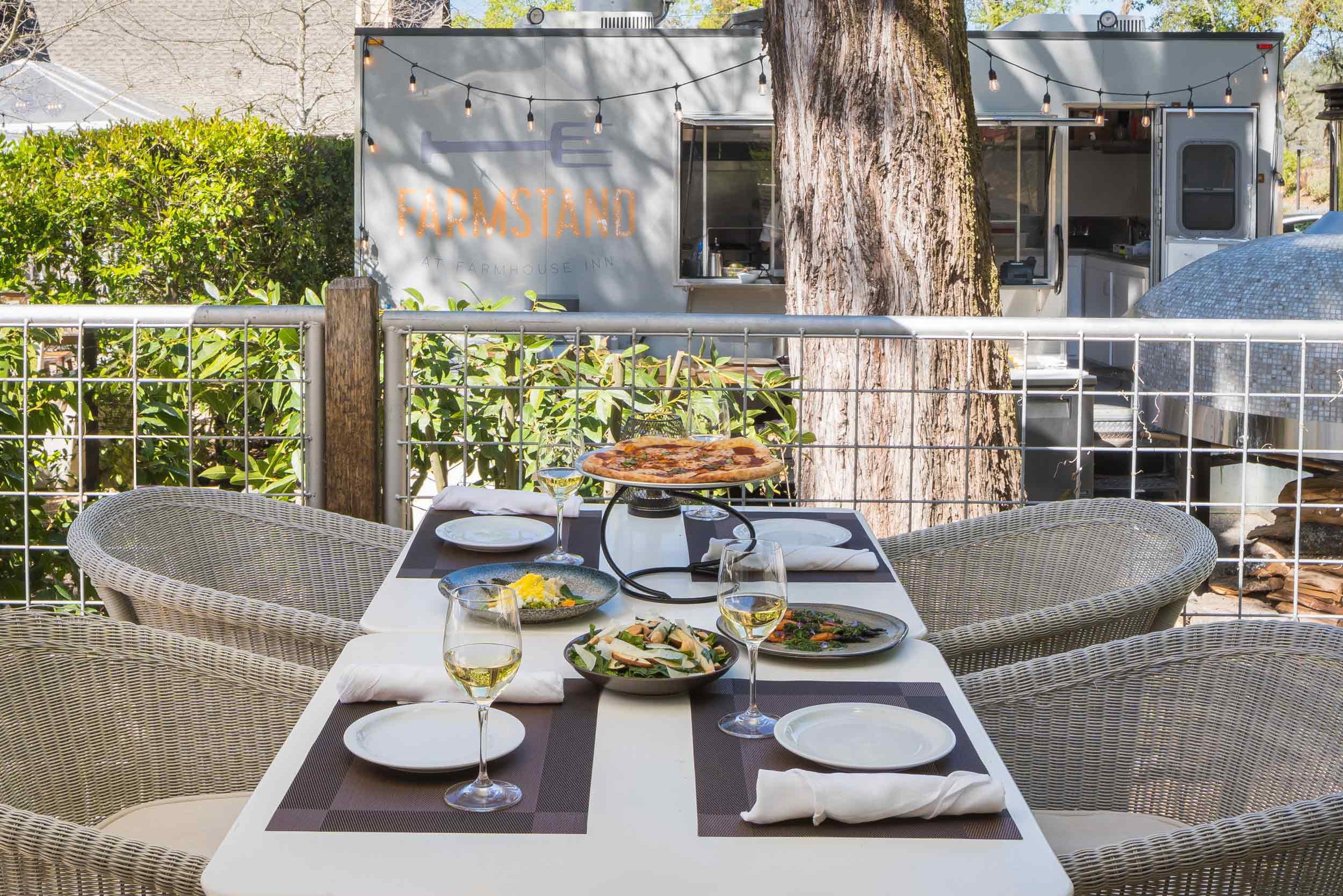 close-up of an outdoor four seated table outside The Farmstand. Four wicker seats, brown placemats, whote plates, two wine glasses along with salad and pizza.