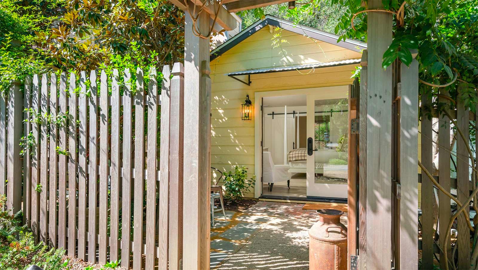 Exterior view of Cottage Room: looking through gate of wood picket fence, yellow exterior of building, beautiful glass double doors. Bed, wood floor and white chair are visible through glass doors.