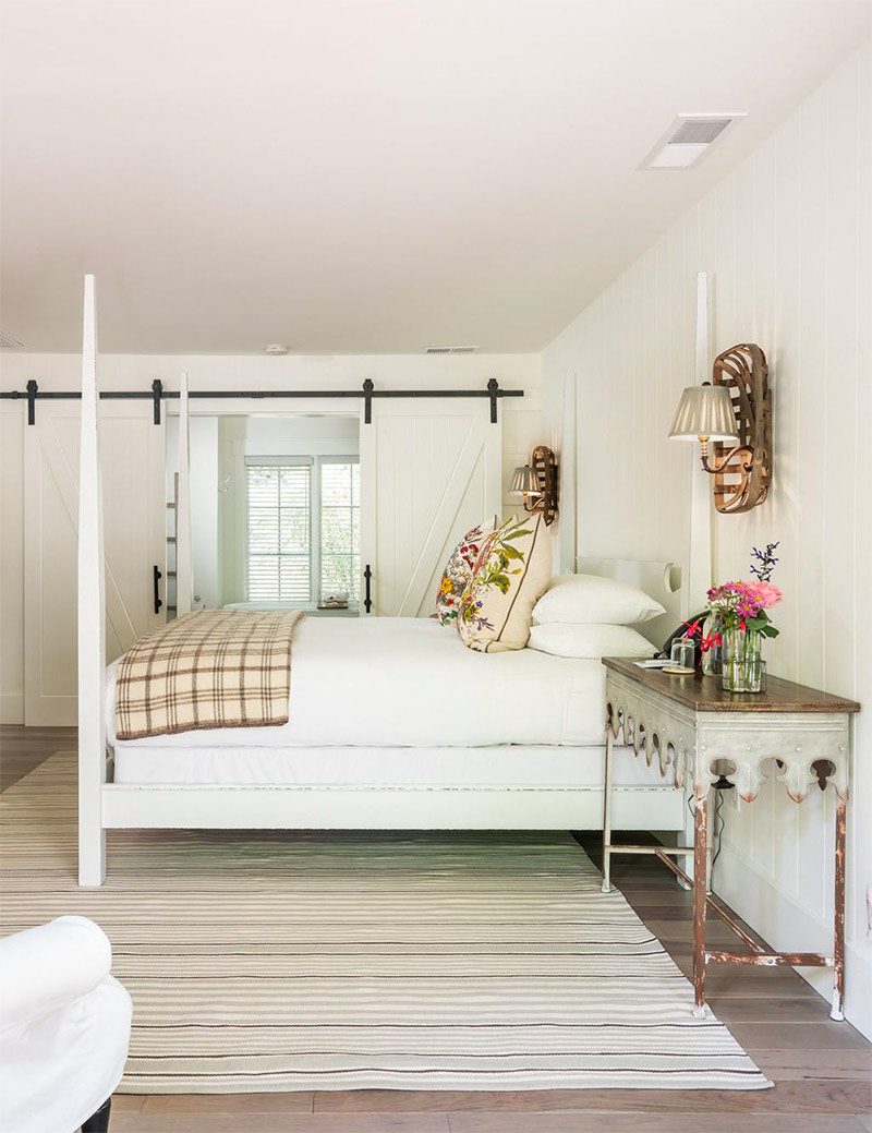 Interior view of hotel room with large bed with white bed posts and white bedding, think area rub under bed, cushioned chairs and elegant table next to bed. Sliding double doors in the back to the bathroom.