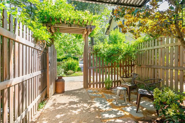Exterior view of Cottage Room patio: tall wood picket fence with path out of a gate and lush greenery in the distance.