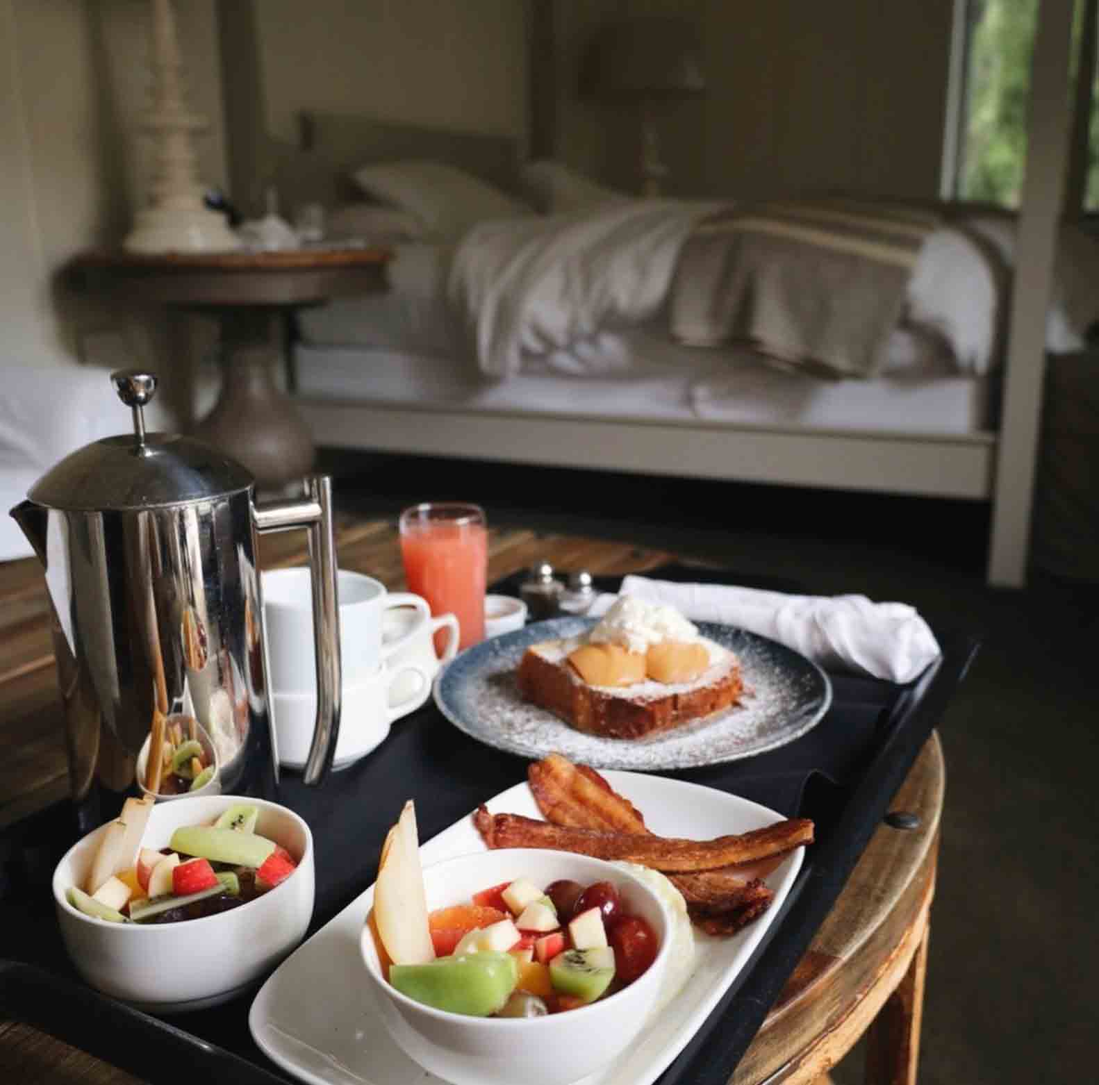 tray of breakfast food and coffee pot on wood table with bed in the background