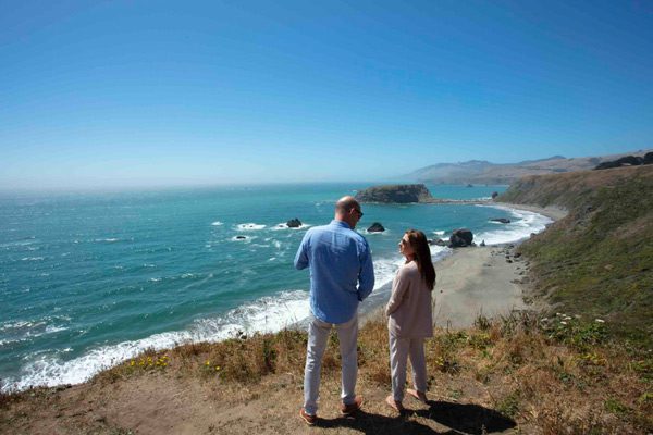 Man and woman on grassy hill overlooking the Pacific Ocean with rolling hills in the distance.