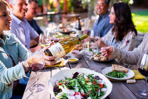 Outdoor gathering people sitting at a wood table with various food items including a green salad and a man pouring Freeman white wine.