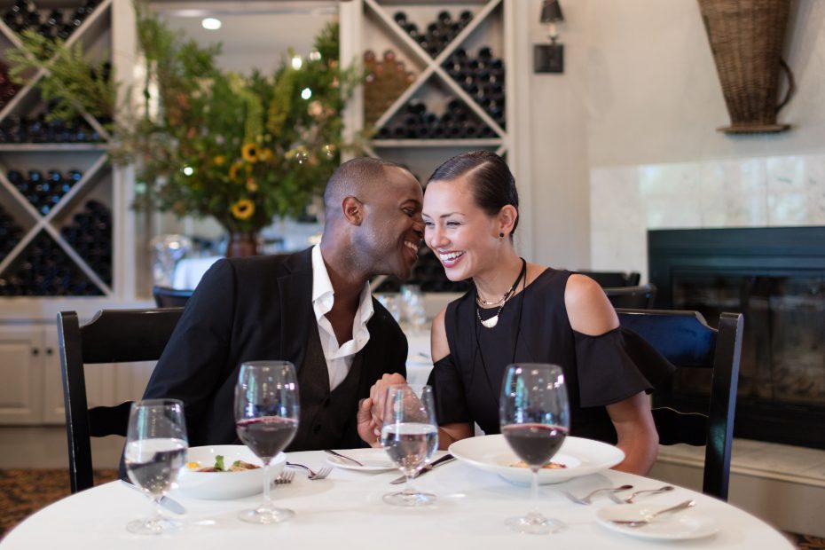 man and woman drinking wine at restaurant and man is whispering in womans ear and laughing