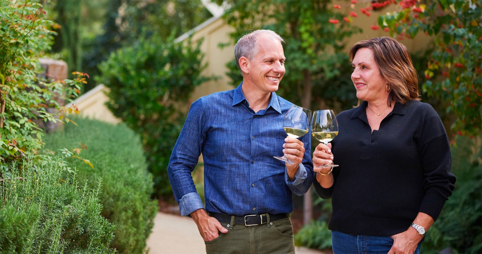 Photo of Joe and Catherine Bartolomei each holding a glass of wine smiling on the property of Farmhouse Inn