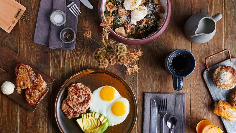 overhead view of breakfast: wood table with various dishes of breakfast including sunny-side-up eggs, avocado, sausage, poached eggs, coffee, milk, fork, spoon and knife.