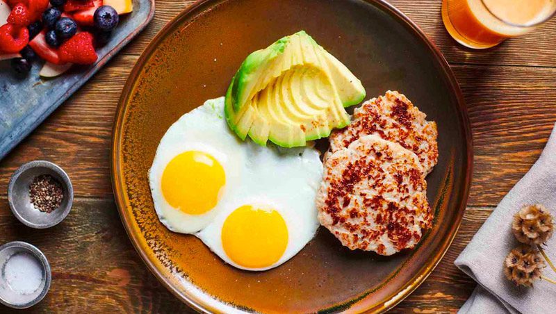 Overhead view of wood table with a breakfast plate: two eggs over-easy, sliced avocado, two sausage patties, juice and fresh berries.