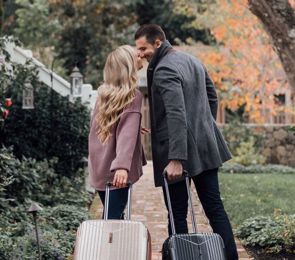 man and woman couple about to kiss , walking down brick path with rolling suitcases, dressed in autumn clothing