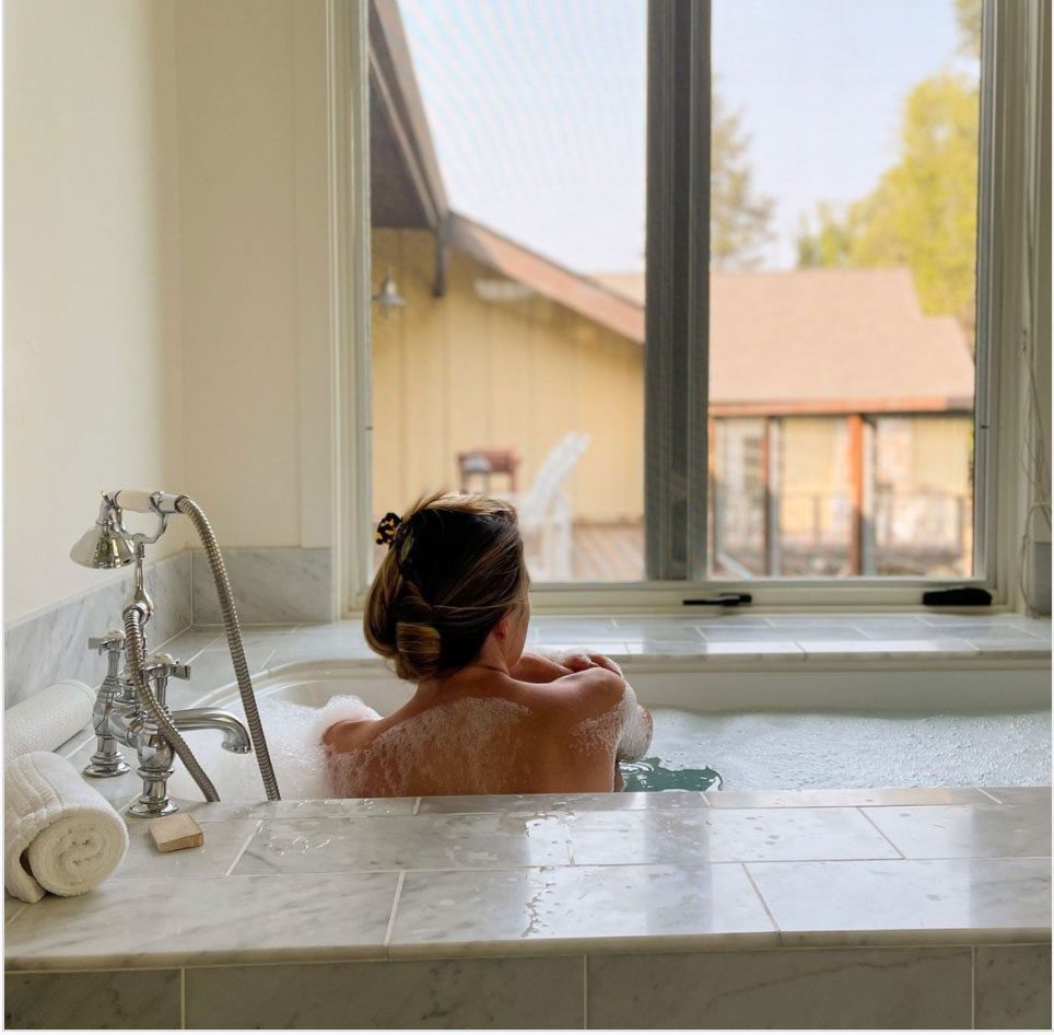 view from behind of the upper back and head of a woman taking a bath, looking out the window