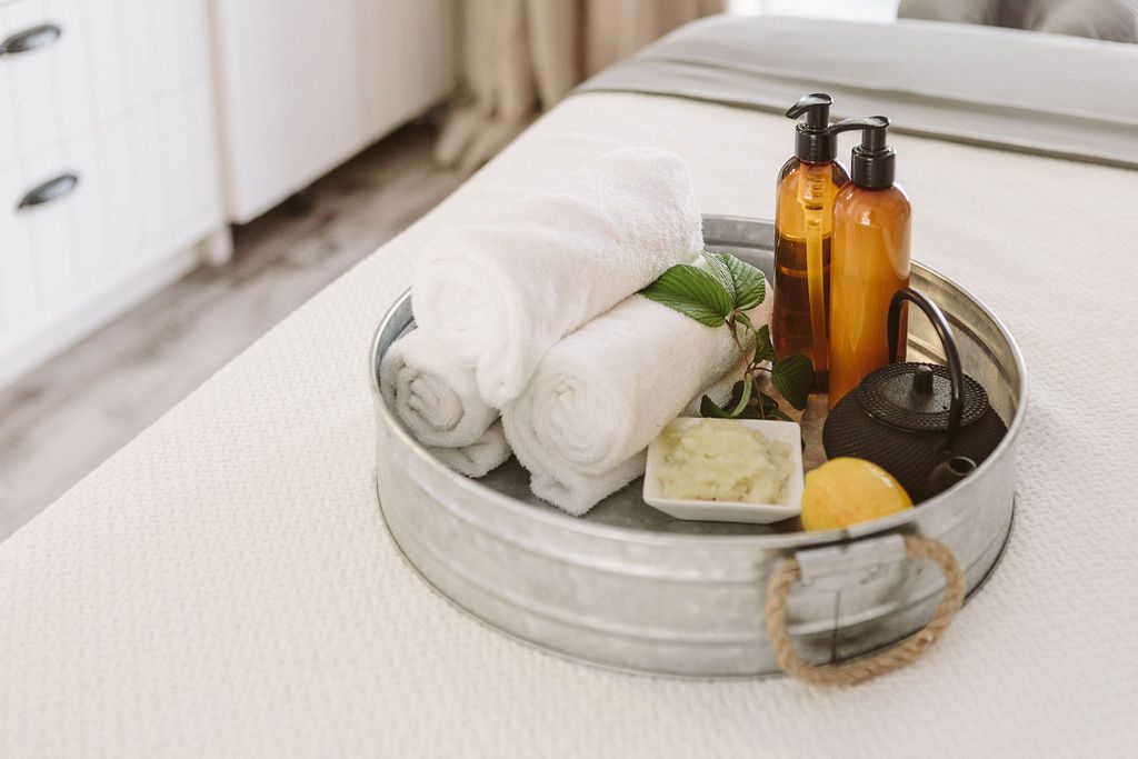 close-up of short metal pail with spa essentials: three rolled towels, two brown pump bottles, small hot water tea kettle, lemon.