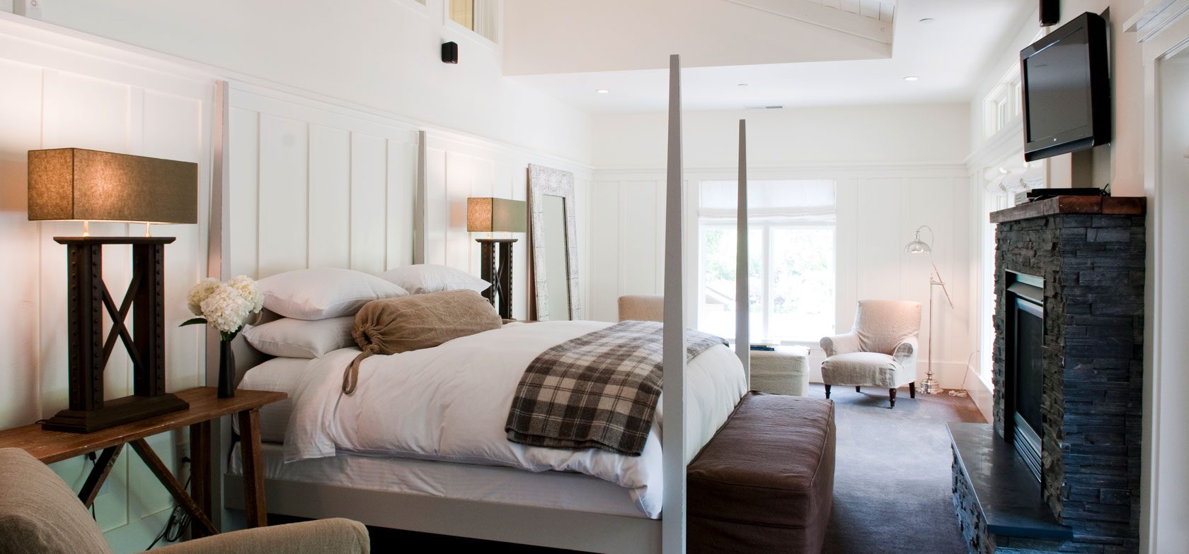 Interior view of Barn One Bedroom Suite. White walls, stone fireplace with mounted TV above, large bed with white bedding with gray bedposts, high ceilings with ceiling fan.