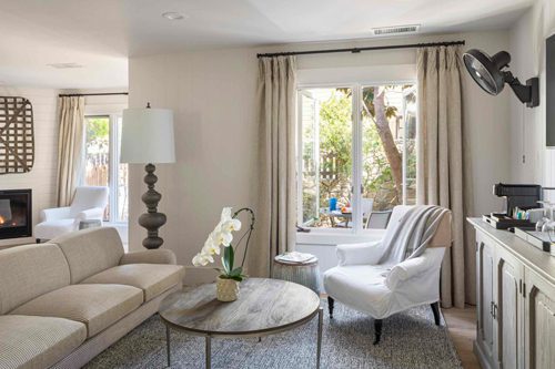 Interior view of Cottage Petite Suite facing large window. Side view of couch wood rounf coffee table, white cushioned chair, area rug, credenza, mid sized flat screen-mounted, large fancy floor lamp, fireplace with fire on.