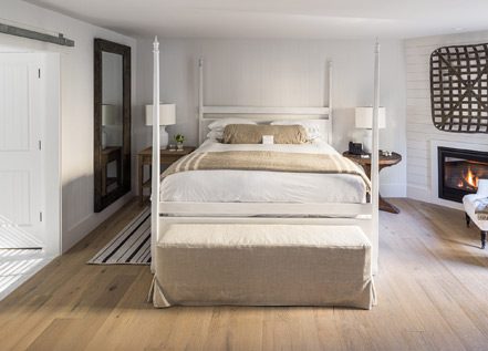 Interior view of Cottage Petite Suite facing bed. Large bed with white bed posts and light bedding, sideview of fireplace with fire, white cushioned chair, and large wall mirror with wood frame. Large paneled wood floors with striped area rug next to bed.