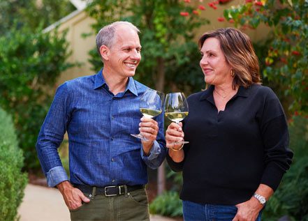 Photo of Joe and Catherine Bartolomei each holding a glass of wine smiling on the property of Farmhouse Inn