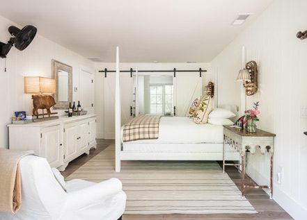 Interior view of hotel room with large bed with white bed posts and white bedding, think area rub under bed, cushioned chairs and elegant table next to bed. Sliding double doors in the back to the bathroom.