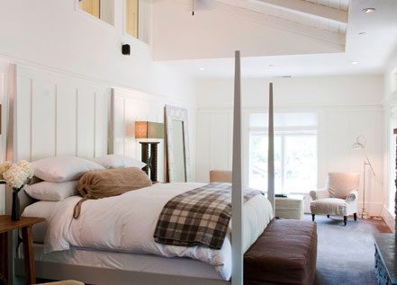 Interior view of Barn One Bedroom Suite. White walls, stone fireplace with mounted TV above, large bed with white bedding with gray bedposts, high ceilings with ceiling fan.