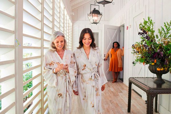 An older woman and younger woman walking happily in matching robes from the Farmhouse Inn Spa. The Spa keeper is in the background.
